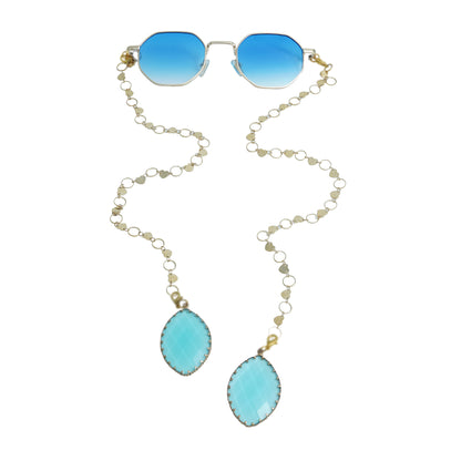 Blue-lensed sunglasses with a gold frame are connected to a delicate gold chain adorned with small circular links, ending in two tear-drop shaped blue charms-front view