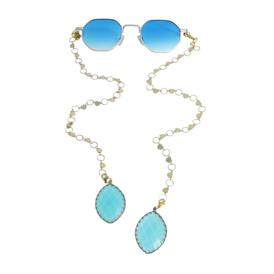 Blue-lensed sunglasses with a gold frame are connected to a delicate gold chain adorned with small circular links, ending in two tear-drop shaped blue charms-front view