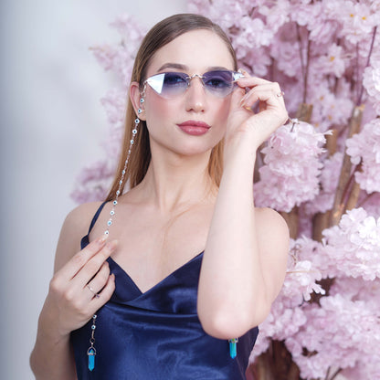 A pair of cat-eye framed sunglasses with a silver frame is connected to a silver evil-eye chain, which holds a pendant shaped blue crystal(Blue Stone)-angle view
