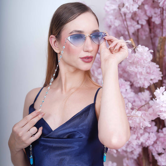 A pair of cat-eye framed sunglasses with a silver frame is connected to a silver evil-eye chain, which holds a pendant shaped blue crystal(Blue Stone)-styling view
